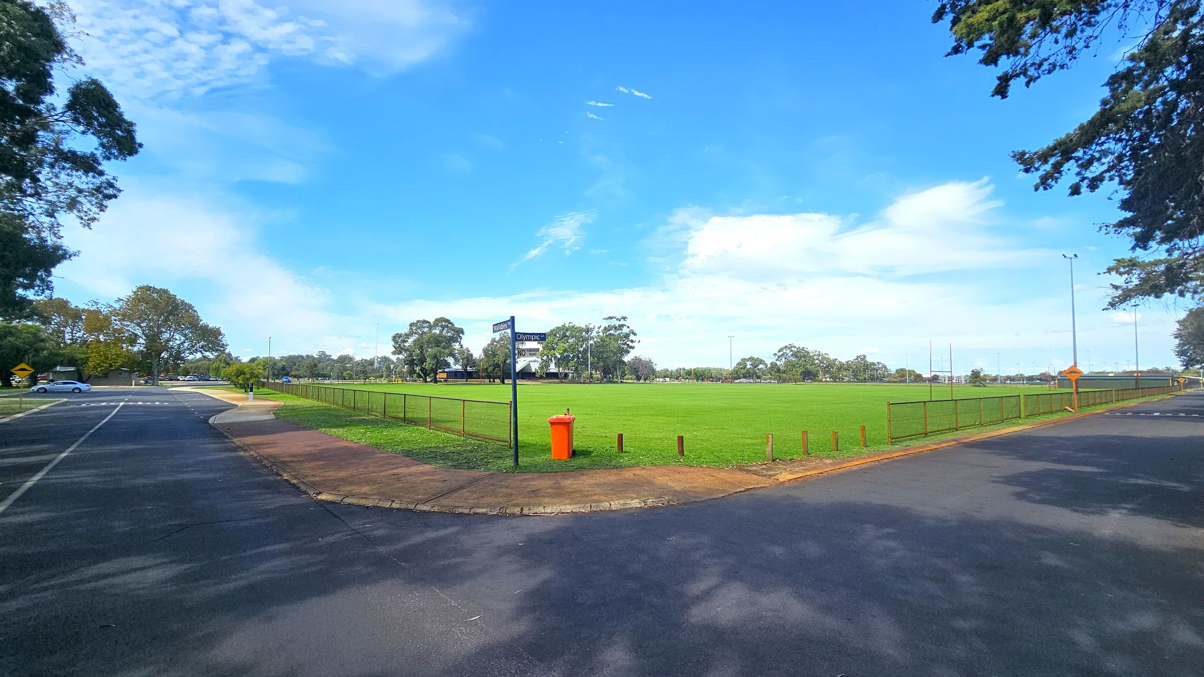 A wide, open grassy field is in the centre, surrounded by a low metal fence. An orange bin is near the fence. To the left, a paved road curves with a few parked cars visible. Trees line the background, providing a natural border. The sky is clear with a few clouds. The foreground shows a paved area with shadows from nearby trees.