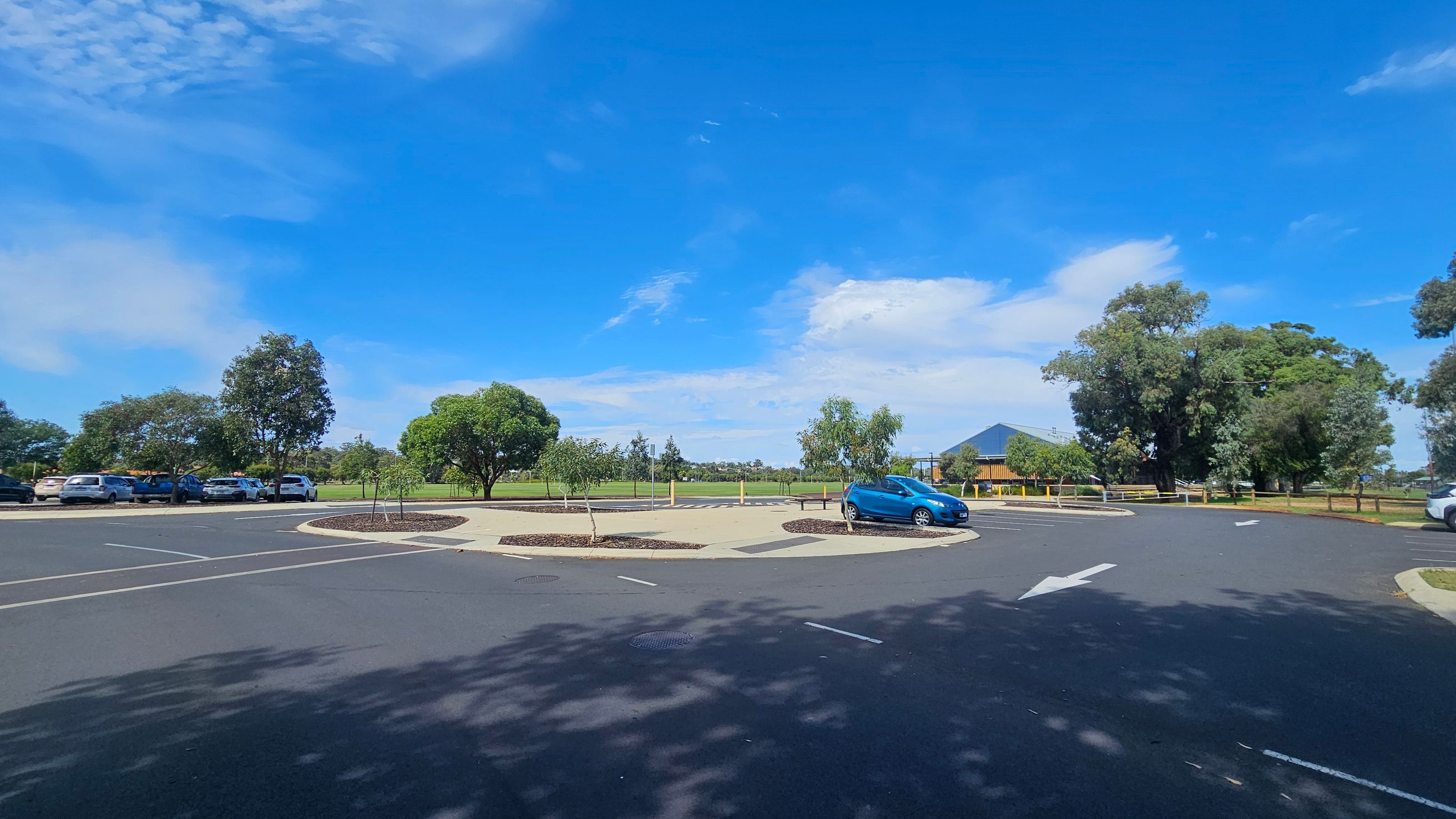 A car park with a roundabout in the centre, featuring a few small trees and a blue car parked on the right. On the left, several cars are parked along the edge, with trees and greenery in the background. A building with a dark roof is visible in the distance on the right. The surface is paved with marked parking spaces and directional arrows.