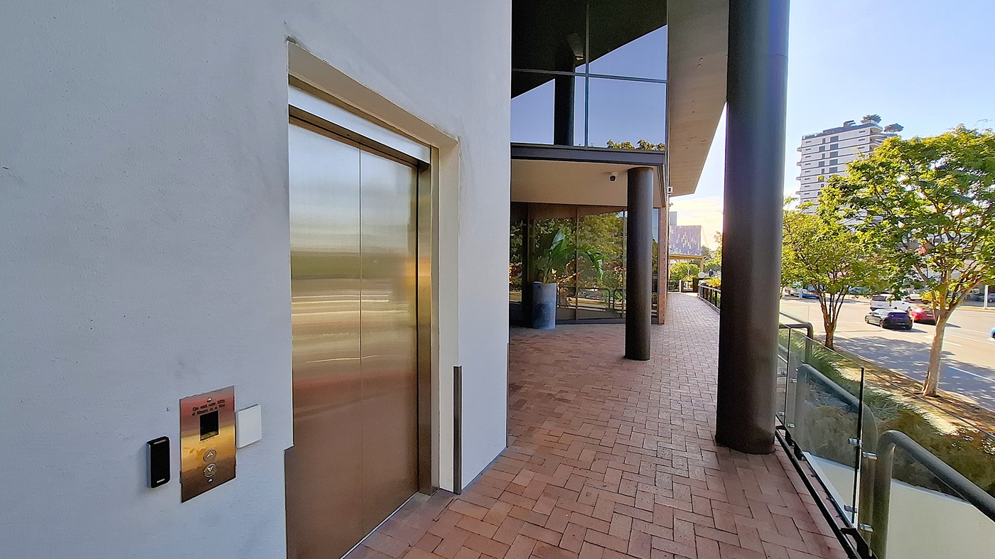 On the left, a stainless steel lift door set in a white wall with a control panel beside it. In the middle, a large black column supports an overhang, with glass windows and a potted plant visible. On the right, a brick walkway bordered by a glass railing, leading to a street with trees and parked cars. The floor is red brick, and the ceiling is white with recessed lighting.