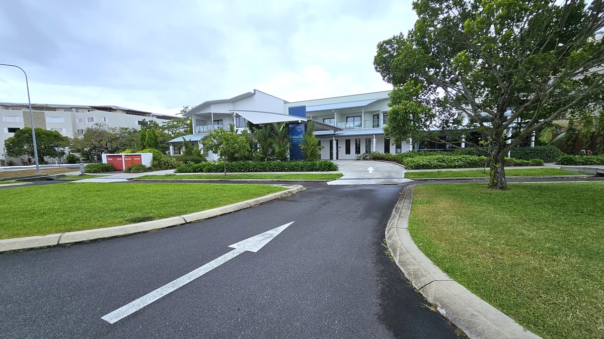 A bitumen road, marked with white arrows, directs towards a concrete pathway to a two-story white building. Ahead, landscaped ornamental plants, manicured grass, and various trees adorn the surroundings. Manicured grass stretches towards the front near the parking area. To the far left, a large white building and tall street lights are visible.
