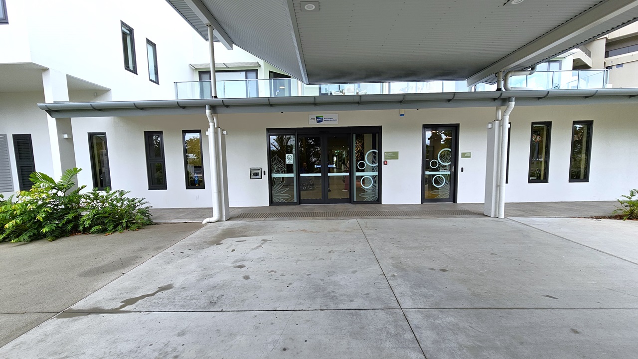 Approaching the building's entrance, a corrugated roof offers shade over the concourse. Ornamental plants adorn the front. Truncated domes mark the floor before the automatic glass sliding door. The building is painted white, with rectangular glass windows. A terrace with a glass railing from the rooftop is also visible.