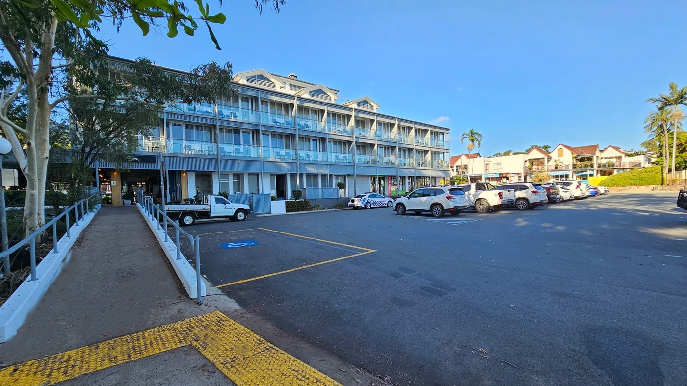 A three-storey building with a modern design, featuring glass balconies and large windows. The ground floor has a chemist shop with a visible sign. Several cars, including a police car with a checkered pattern, are parked in front. The building is surrounded by small shrubs and plants. The sky is clear and blue.