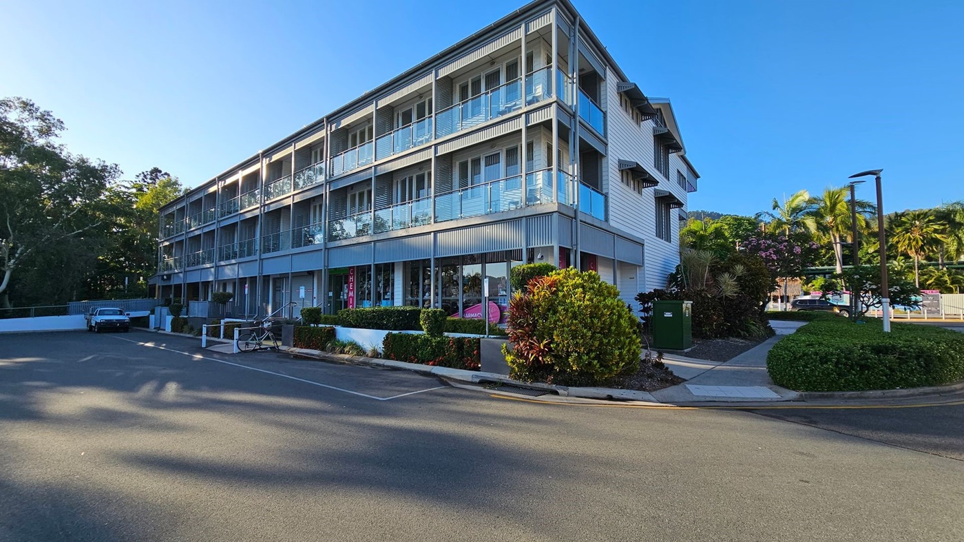 A three-storey building with a modern design featuring glass balconies and a grey facade. On the ground floor, there are shopfronts with signs, including one for a chemist. In the foreground, a paved road leads to a parking area with a parked car and a bicycle. The right side has landscaped greenery with bushes and palm trees. The background includes more trees and a clear blue sky.