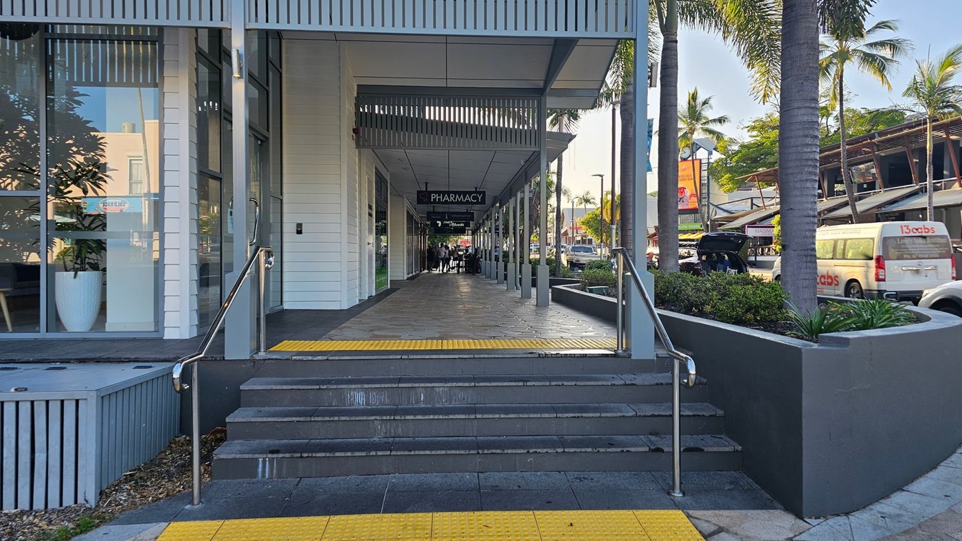A walkway leading to a pharmacy, with steps and a ramp in the foreground. The left side features large glass windows with a potted plant inside. The right side has a raised garden bed with palm trees and a parked van. The ceiling is covered with slatted panels, and the floor is tiled. In the background, there are more shops and people walking.