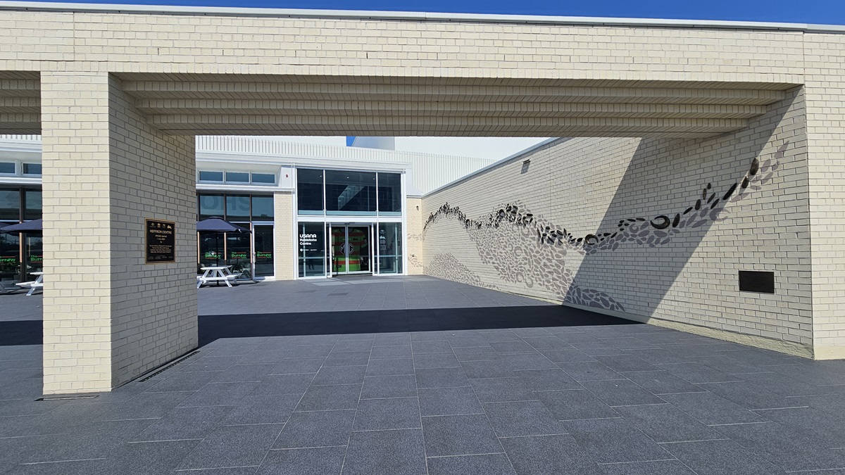 The building has a generous front with a brick wall facade featuring intricate design. A plaque displaying facility information is mounted on the left wall. Behind it is a two-story modern structure with glass walls and corrugated roofing. Tables and chairs are arranged under large umbrella shades.