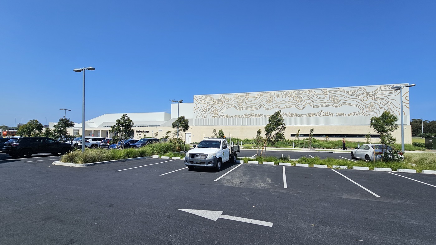 Next to the facility is the car park, featuring line markings and occupied by a few cars. On the left, there's a concrete plant box with trees and grass. Adjacent to it is the building, presenting a brick wall facade with intricate design and the facility name. Street lights are visible.