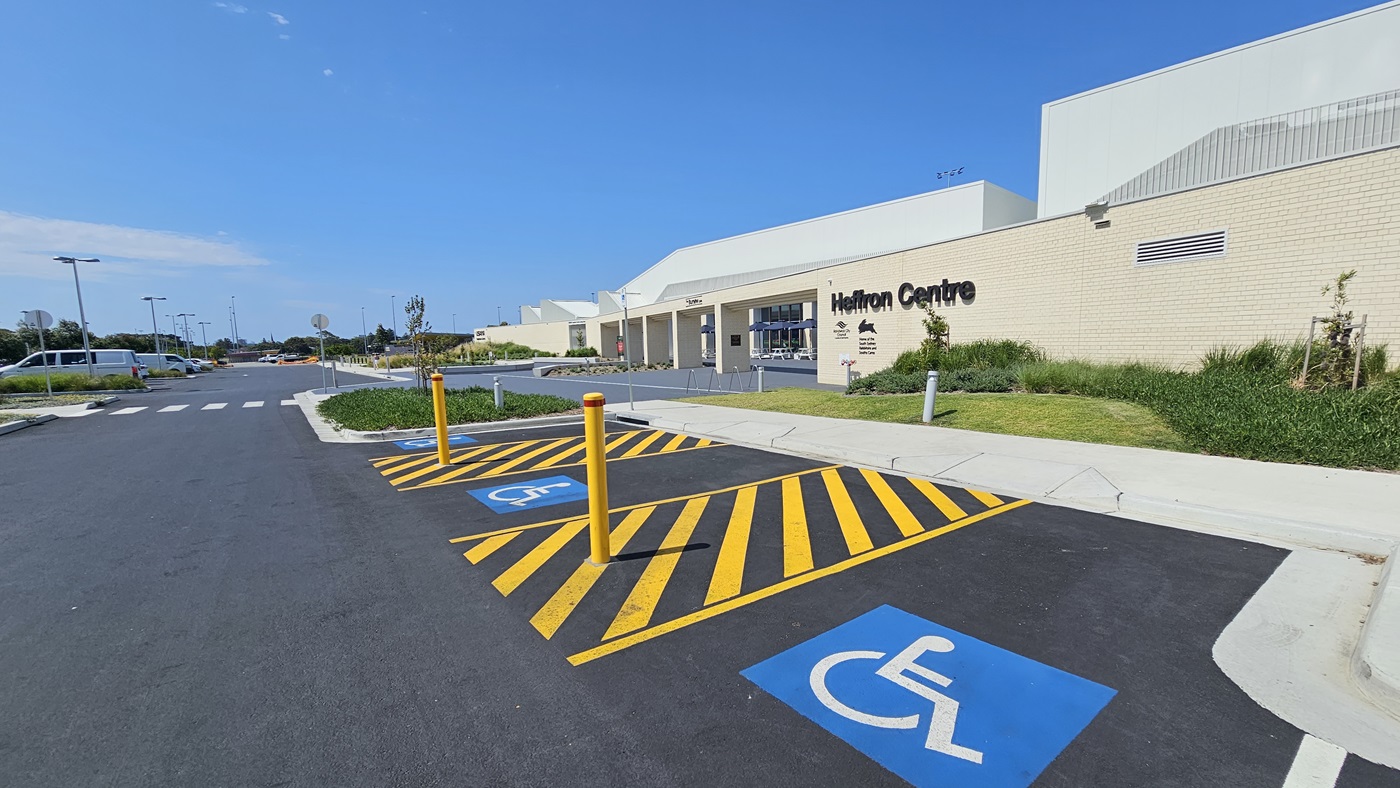 The building features a spacious front with a brick wall facade, showcasing intricate design and the facility name. To the right, there are accessible car parking spaces marked by blue wheelchair signs, yellow bollards, and yellow floor markings. In the middle, a pedestrian lane and concrete pathway run between two landscapes, extending towards the building. Street signs and street lights are visible.