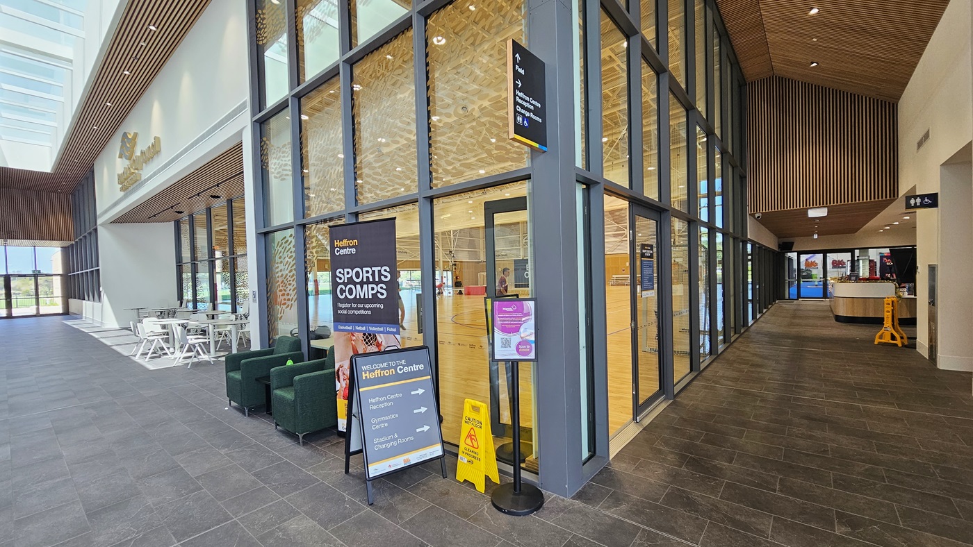 To the left, tables and chairs are arranged. The glass wall features a golden logo and small leaf-like designs in gold colors, with free-standing banners positioned on the wall. To the right is a corridor leading to the Heffron Centre reception area. The floors have matte grey tiles, and the ceiling features intricately designed wooden panels with LED lights. Further right, the reception desk and the gymnastics area are visible.