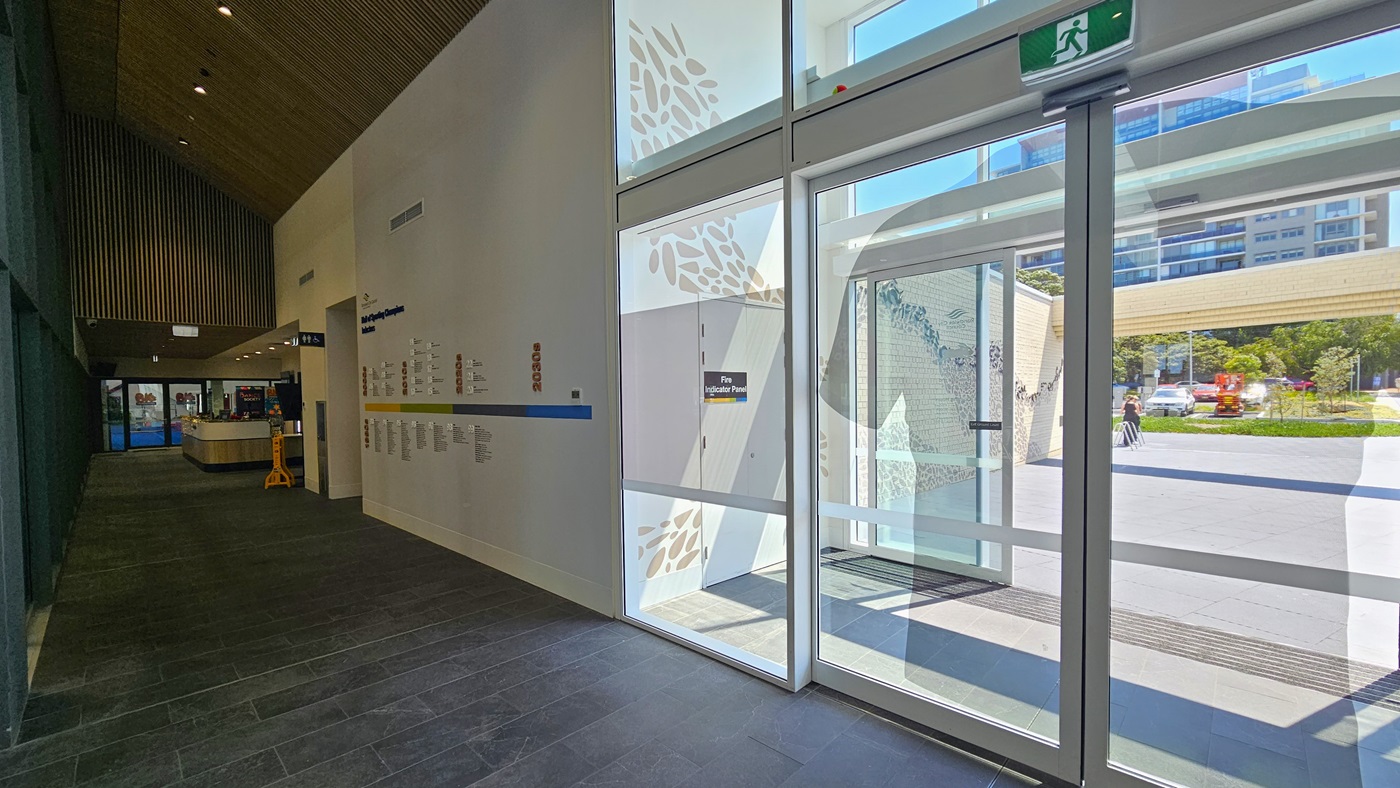 The corridor leads to the Heffron Centre reception desk. The right wall features a 'Hall of Sporting Champion Inductees.' Straight ahead is the reception desk and the gymnastic area. To the right is the entrance to the building featuring double automatic glass doors. The wall outside features intricate designs. Outside, the car park, tall commercial buildings, and trees are visible.