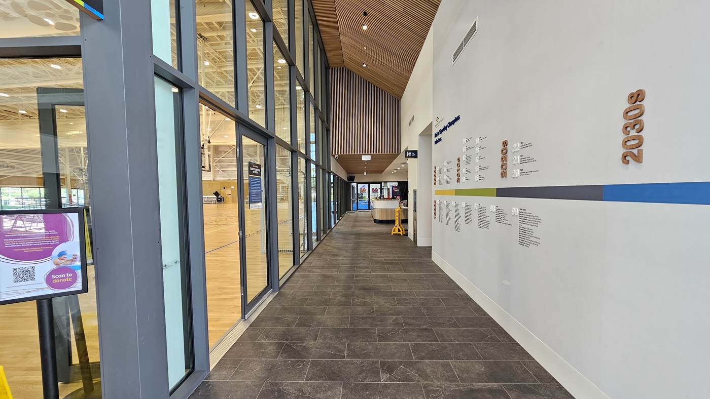 The corridor directs to the Heffron Centre reception desk. The right wall features a 'Hall of Sporting Champion Inductees.' Straight ahead is the reception desk and the gymnastics area. To the left is the sports stadium with timber floors and an industrial ceiling. Posters are affixed to the glass wall. The corridor has grey tiles, and the ceiling features an intricate wooden design.