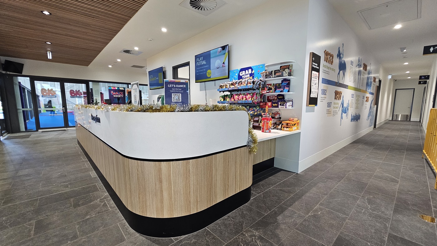 The reception desk displays a white and timber design front with food items, banners, posters, and garlands. Two flat-screen TVs are mounted on the wall. The right corridor has a white wall painted with historical facts, and accessible bathroom signage is mounted on the walls to the right. To the left is the gymnastics area. The corridor has grey tiles, and the ceiling features an intricate wooden design.