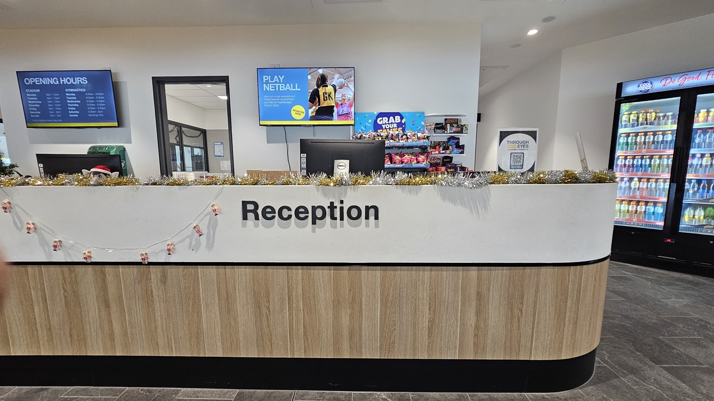 The reception desk has a white and timber design front, displaying food items, banners, posters, and garlands. Two computers are placed on top of the desk. In the middle, a door leads to the staff office. Two flat-screen TVs are mounted on the wall. To the right, a vending machine offers cold drinks. The floor has grey tiles, and the ceiling is all white with LED lights.