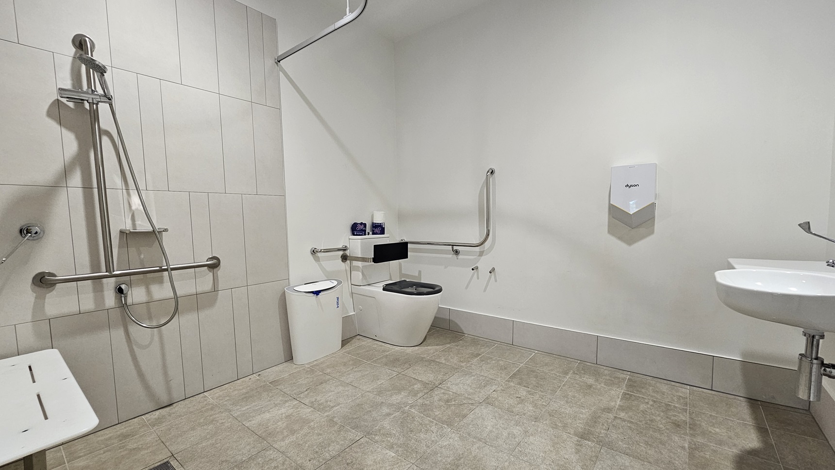 Inside the accessible bathroom, to the left corner is a toilet bowl equipped with hand grabs, accompanied by a rubbish bin. Adjacent is a shower with hand grabs and a placeholder for a shower curtain. A foldable bench chair is positioned to its left. A washbasin and a tissue dispenser are mounted on the right wall. The floor features grey tiles, and the wall is all white.