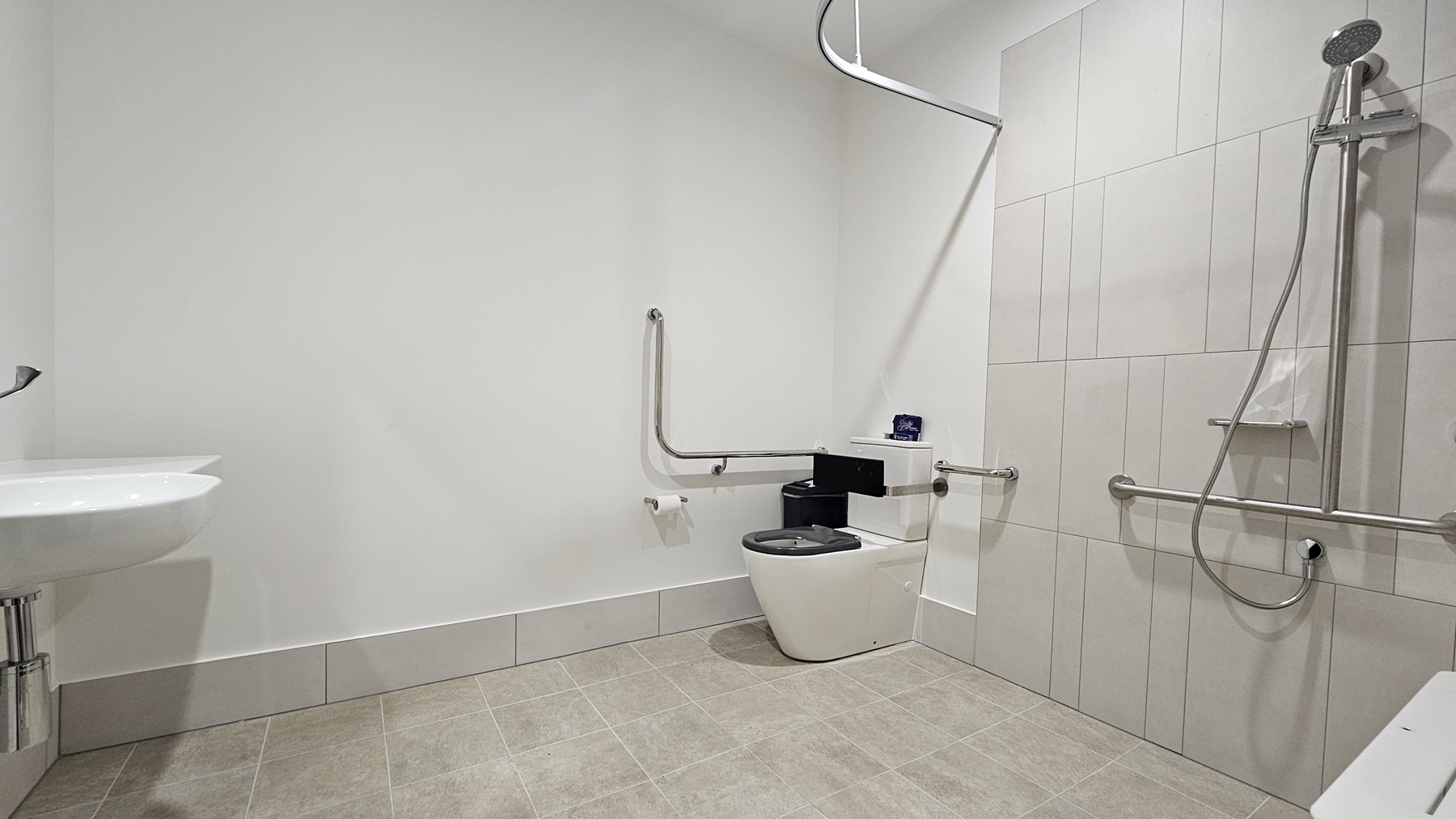 Inside the accessible bathroom, to the right corner is a toilet bowl equipped with hand grabs, accompanied by a rubbish bin. Adjacent is a shower with hand grabs and a placeholder for a shower curtain. A foldable bench chair is positioned to its right. A washbasin and a tissue dispenser are mounted on the left wall. The floor features grey tiles, and the wall is all white.