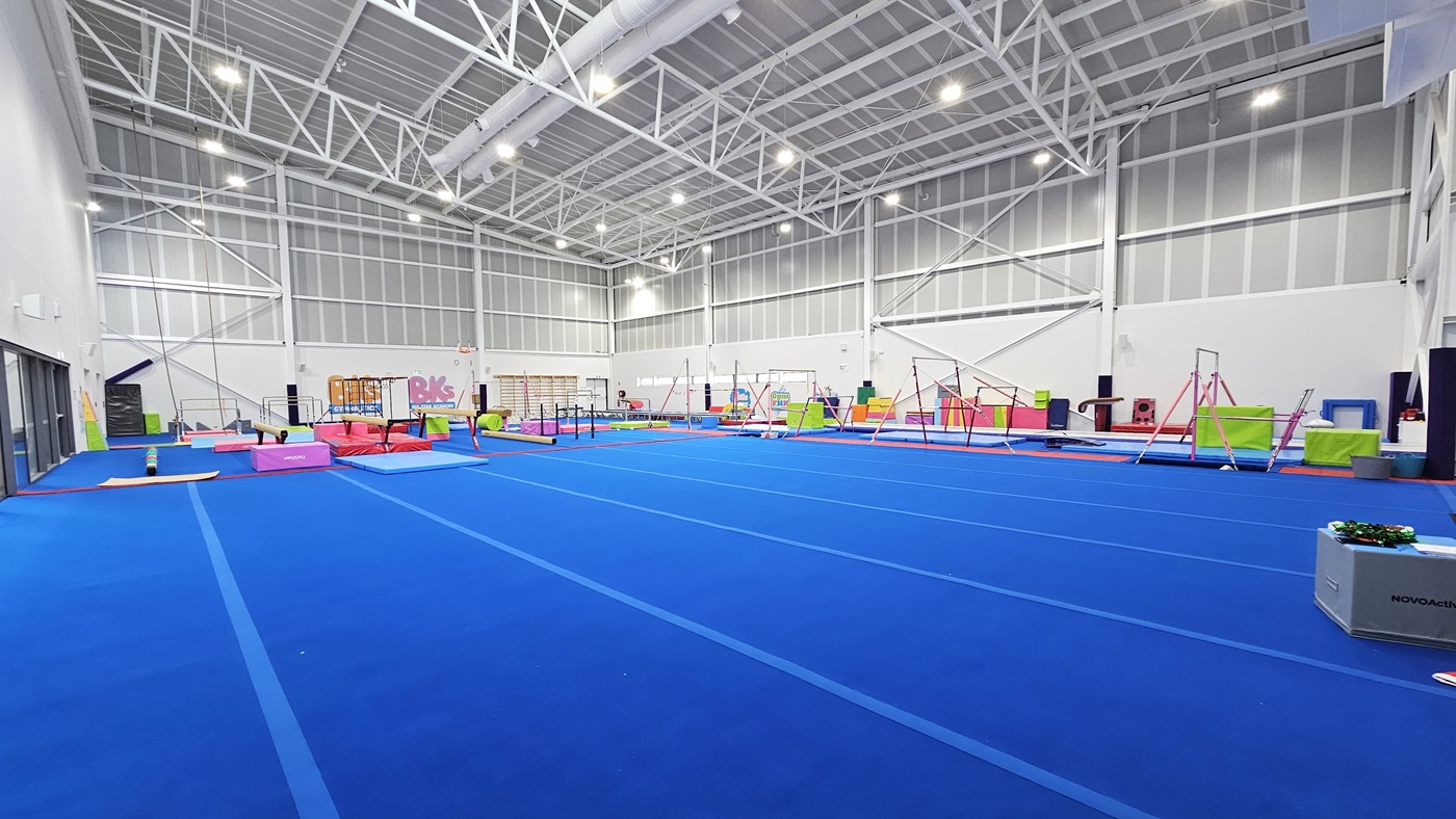 The entrance to the Gymnastics area is through a manual outward-opening glass door, equipped with "BK Gymnastics" stickers. A non-slippery mat is placed on the floor. To the right is a retractable banner and yellow and red rubbish bins. To the left is the glass door entrance to the Sports Stadium, also equipped with a poster. A flat-screen TV is mounted to the upper right wall. Inside the facility, bright blue foam is visible.