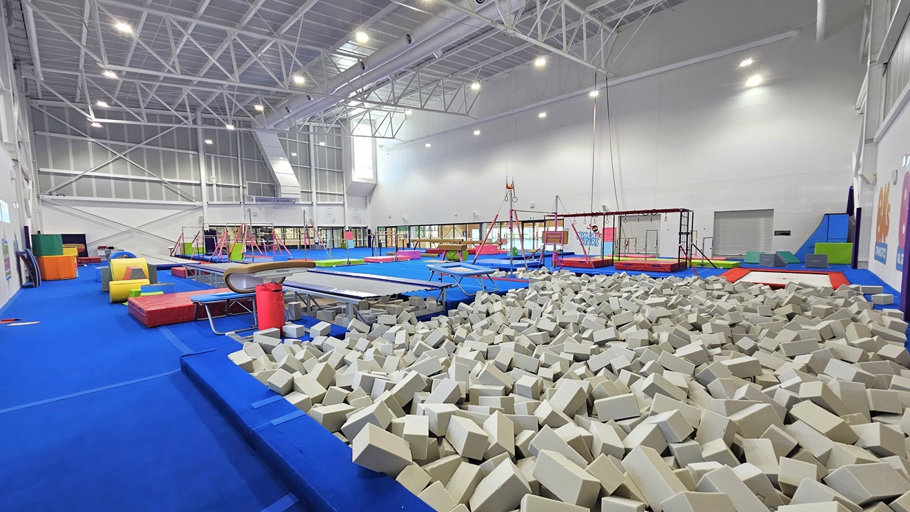 The floor is made of blue bouncy foam. The left and right walls feature white with paintings of the facility's name. Various gymnastics equipment such as balance beams, single bar trainers, horizontal bars, vault springboards, mats, and pommel horses are available. Brightly colored trampolines and tumbling blocks are also present. A foam pit with grey foams is positioned near the wall on the right. The high ceiling features an industrial look with exposed white metal beams.