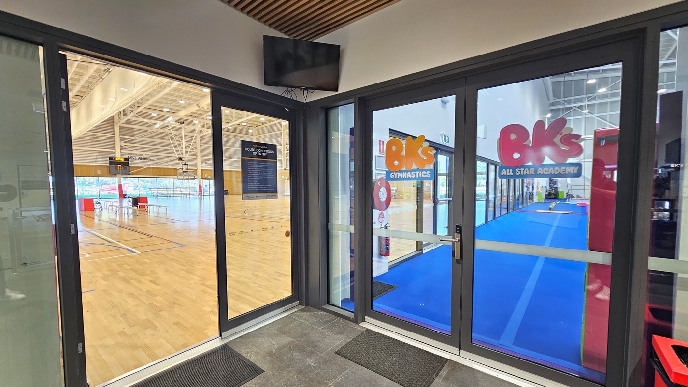The entrance to the Sports Stadium is through a manual inward-opening glass sliding door. A poster is affixed to the wall. Non-slippery mats are placed on the flooring. To the right is a glass door entrance to BK's Gymnastics, equipped with stickers. A flat-screen TV is mounted in the upper corner. Inside the Stadium, the timber floor and the industrial-designed ceiling are visible.