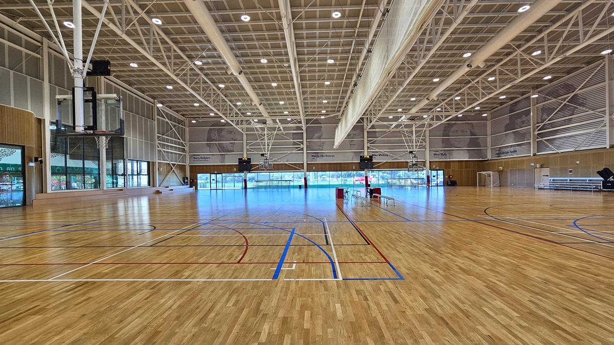 Inside are two basketball courts with timber floors, marked for various ball games. The stadium features timber floors and an industrial ceiling with exposed white beams, LED lights, and air conditioning vents. A screen net is positioned on the ceiling, serving as a separator between the two courts. Tables and chairs are arranged in the middle of the two courts. Distinctive paintings of sports legends adorn the upper wall. On the left is tiered timber seating, and on the right is tiered seating.