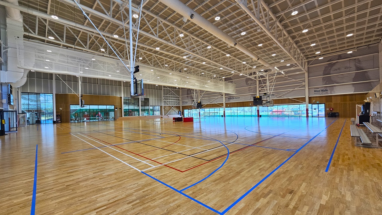 Inside, there are two basketball courts with timber floors, marked for various ball games. The stadium features timber floors and an industrial ceiling with exposed white beams, LED lights, and air conditioning vents. A screen net is positioned on the ceiling, serving as a separator between the two courts. Tables and chairs are arranged in the middle of the two courts. Distinctive paintings of sports legends adorn the upper wall. Tiered seating is visible.