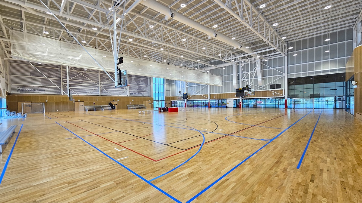 Inside, there are two basketball courts with timber floors, marked for various ball games. The stadium features timber floors and an industrial ceiling with exposed white beams, LED lights, and air conditioning vents. A screen net is positioned on the ceiling, serving as a separator between the two courts. Tables and chairs are arranged in the middle of the two courts. Distinctive paintings of sports legends adorn the upper left wall. Tiered seating is visible on the left.