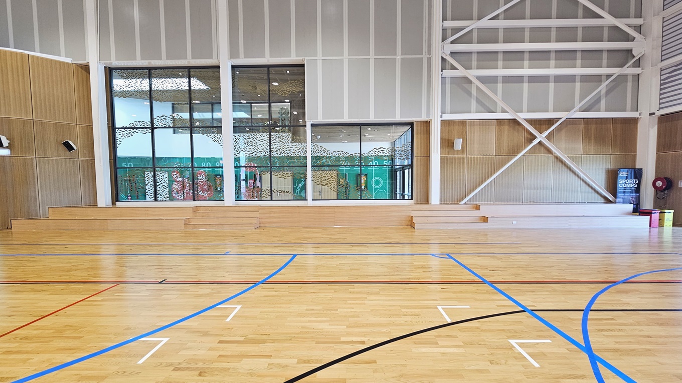 The stadium features timber floors marked for various ball games. The left and right walls have timber designs with mounted audio speakers. Rubbish bins and a fire hose reel, along with a retractable banner, are placed in the right corner. The middle glass features a foliage-shaped design in gold. Below it is a tiered timber seating.