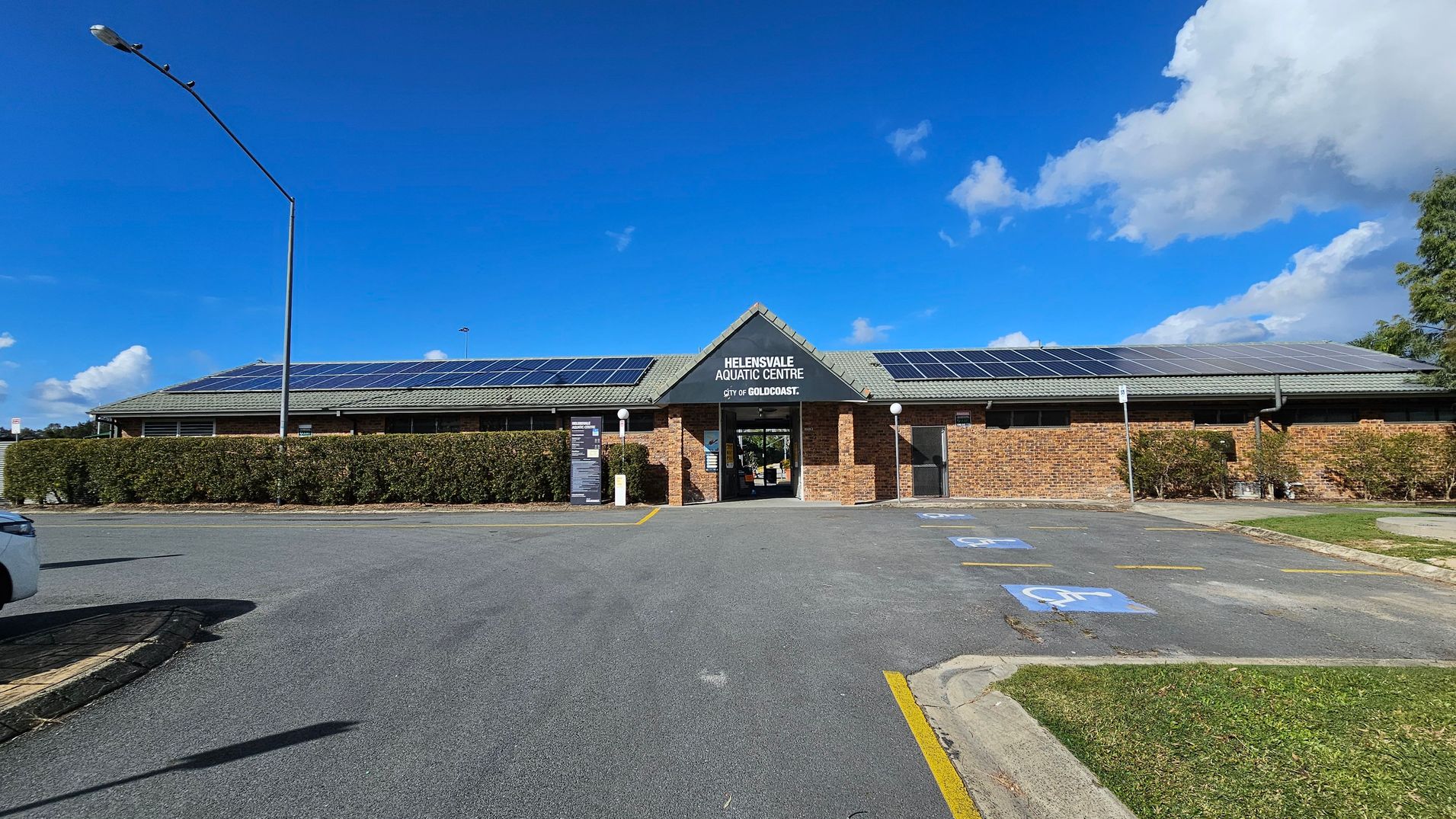 A single-storey brick building with a grey roof featuring solar panels. The entrance is centrally located with a sign above it. A hedge runs along the front of the building. The foreground shows a car park with marked disabled parking spaces and a streetlamp on the left. A patch of grass is visible on the right.