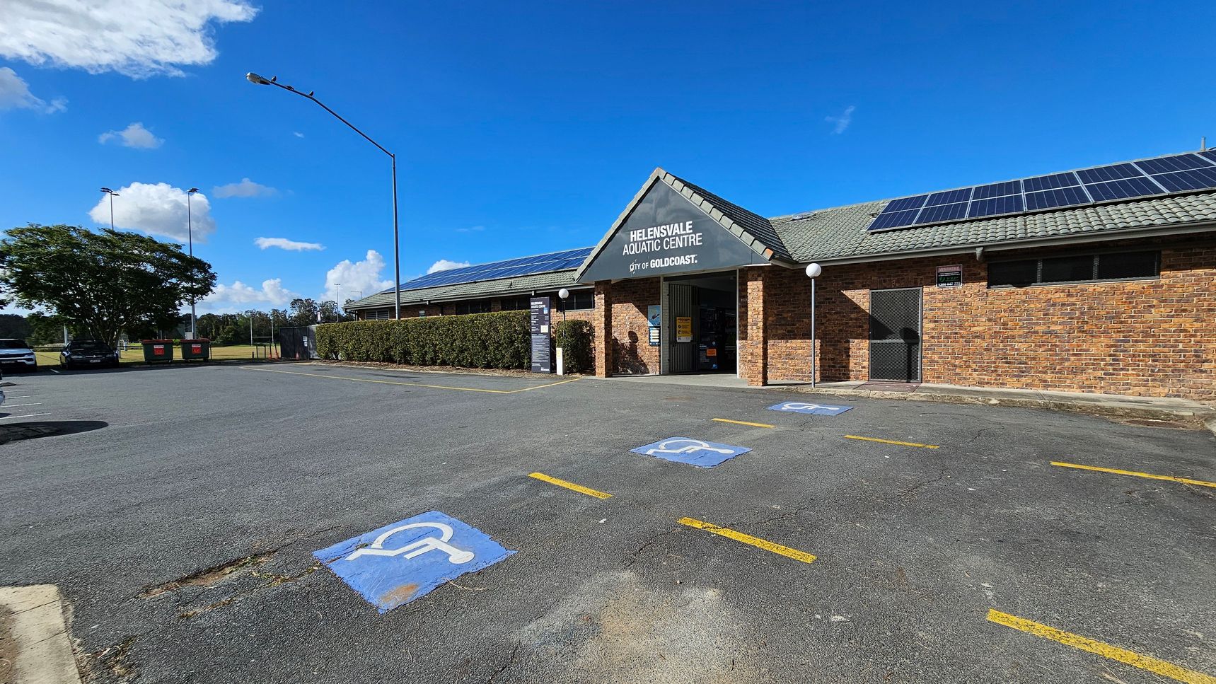 A brick building with a grey tiled roof and solar panels on top. The entrance is marked "Helensvale Aquatic Centre" with a sign for the City of Gold Coast. In front, there are several parking spaces with blue wheelchair symbols. A hedge runs along the side of the building. On the left, a tree and some parked cars are visible. The sky is clear and blue, and there are streetlights in the background. The ground is paved with some patches of grass.
