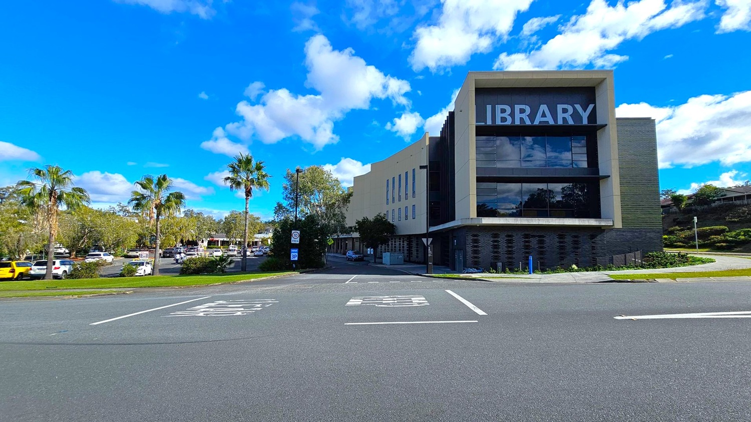 A modern library building is on the right, with large windows and the word "LIBRARY" prominently displayed. The foreground features a road with marked parking spaces. On the left, there are several palm trees and a parking area filled with cars. The background includes more trees and a clear blue sky with scattered clouds.