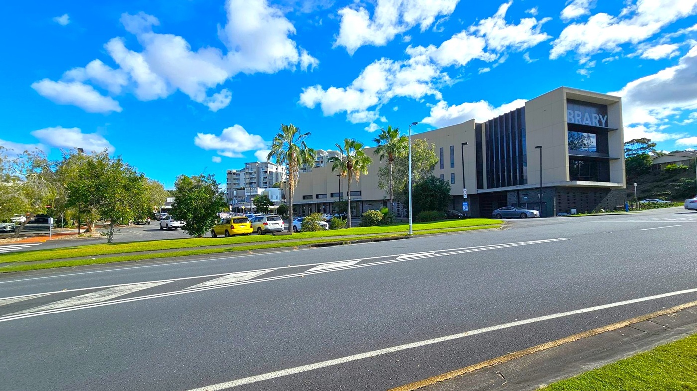 A modern building with the word "BRARY" visible on the right side, likely part of a library sign. The structure is surrounded by palm trees and other greenery. Several cars are parked in a lot to the left, including a yellow one. The foreground features a multi-lane road with white markings. In the background, there are additional buildings and more trees under a blue sky with scattered clouds.