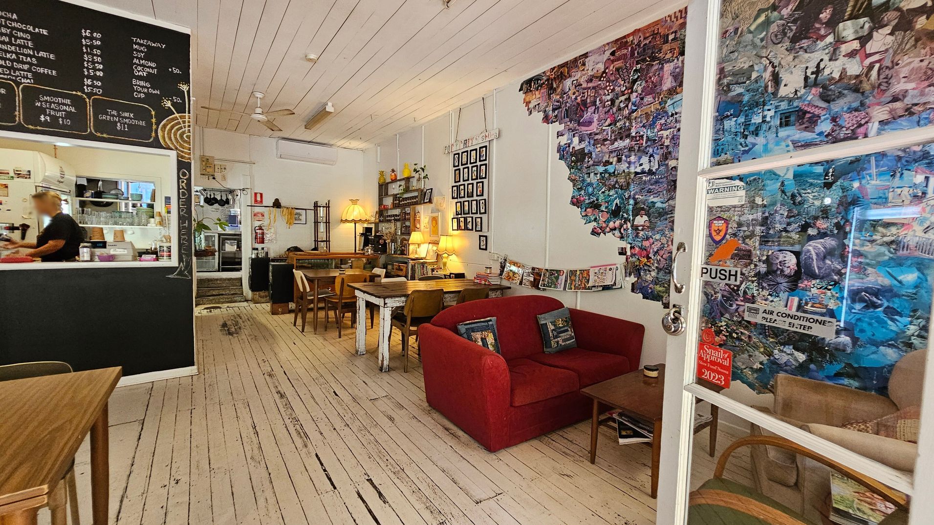 A cosy café interior with a rustic wooden floor and white panelled ceiling. On the left, a blackboard menu is displayed above a counter where a person is working. In the centre, wooden tables and chairs are arranged, with a vintage lamp and shelves in the background. On the right, a red sofa and a collage-covered wall add a colourful touch. A glass door with stickers and a "PUSH" sign is visible, leading to another seating area.