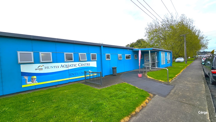 A closer look at the entrance leading to the Huntly Aquatic Centre. A dark grey pathway with steel handrails connects the sidewalk to the main glass entrance doors of the center. Small square glass windows are placed alongside the front wall of the center. Cars are parked in front, and to the back is a white car. The landscape of the area is well maintained as grasses are at the same height and can be distinguished by a yellow marking on the side.