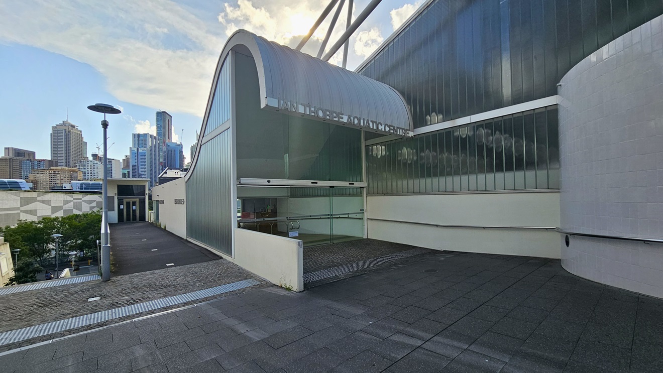 The building showcases a distinctive corrugated wave roofing design in blue. The left wall features glass structures, and a glass door entry is on the left. The facility name is written on top of the wave-shaped roofing. The entrance has an automatic glass sliding door with truncated domes. To the left is a pathway leading to another entrance, overlooking the section below. Tall light posts are present. Further ahead, tall commercial buildings come into view.