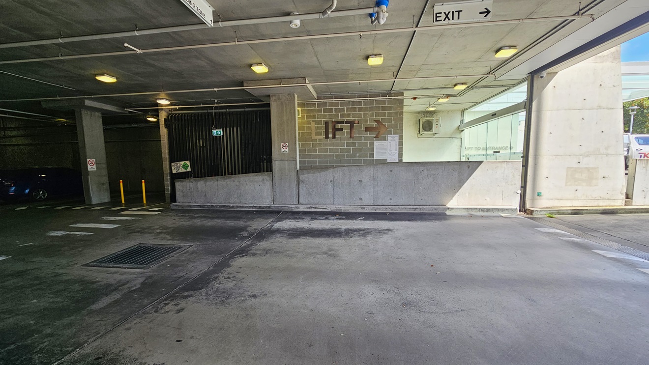 The underground parking includes a pedestrian lane on the left, marked by yellow bollards. The middle brick wall has a sign indicating the lift direction. The low ceiling features directional signs, LED lights, and exposed pipes. Safety signs are affixed to the wall, and a visible water drainage system is present.