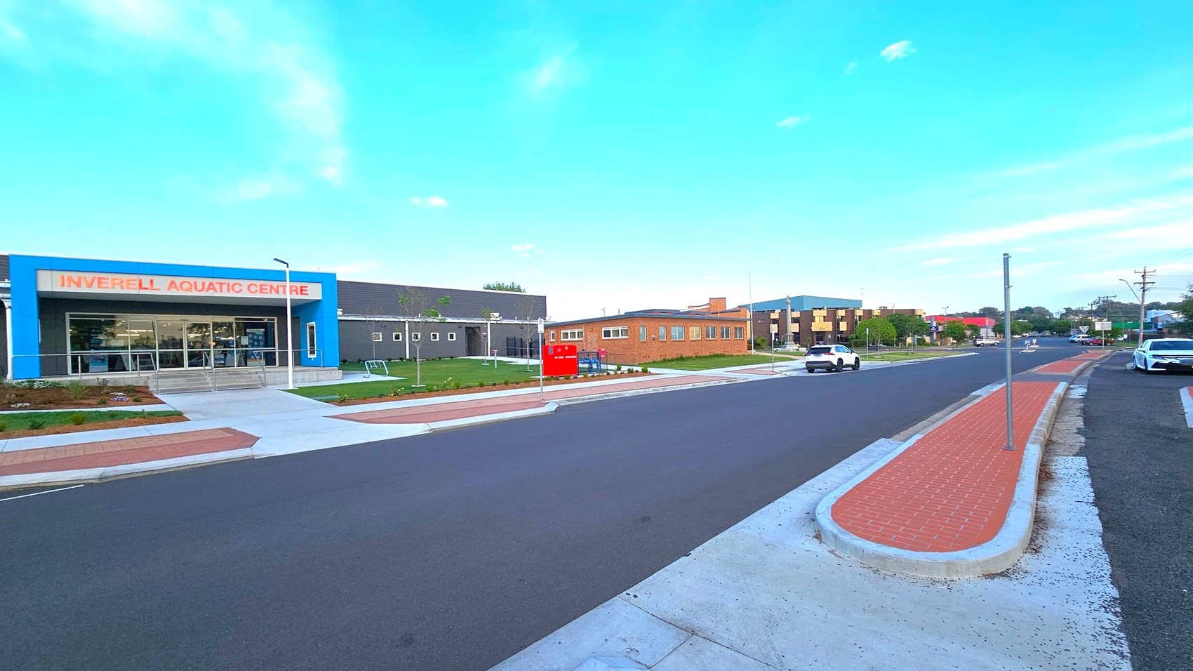 On the left, a building with a blue facade and large windows is labelled "Inverell Aquatic Centre". In the middle, a wide road with a red brick pedestrian island runs parallel to the building. On the right, a white car is parked along the kerb, and another vehicle is driving down the road. The background features a mix of modern and older buildings, with some greenery visible. The pavement is concrete, and the sky is clear.