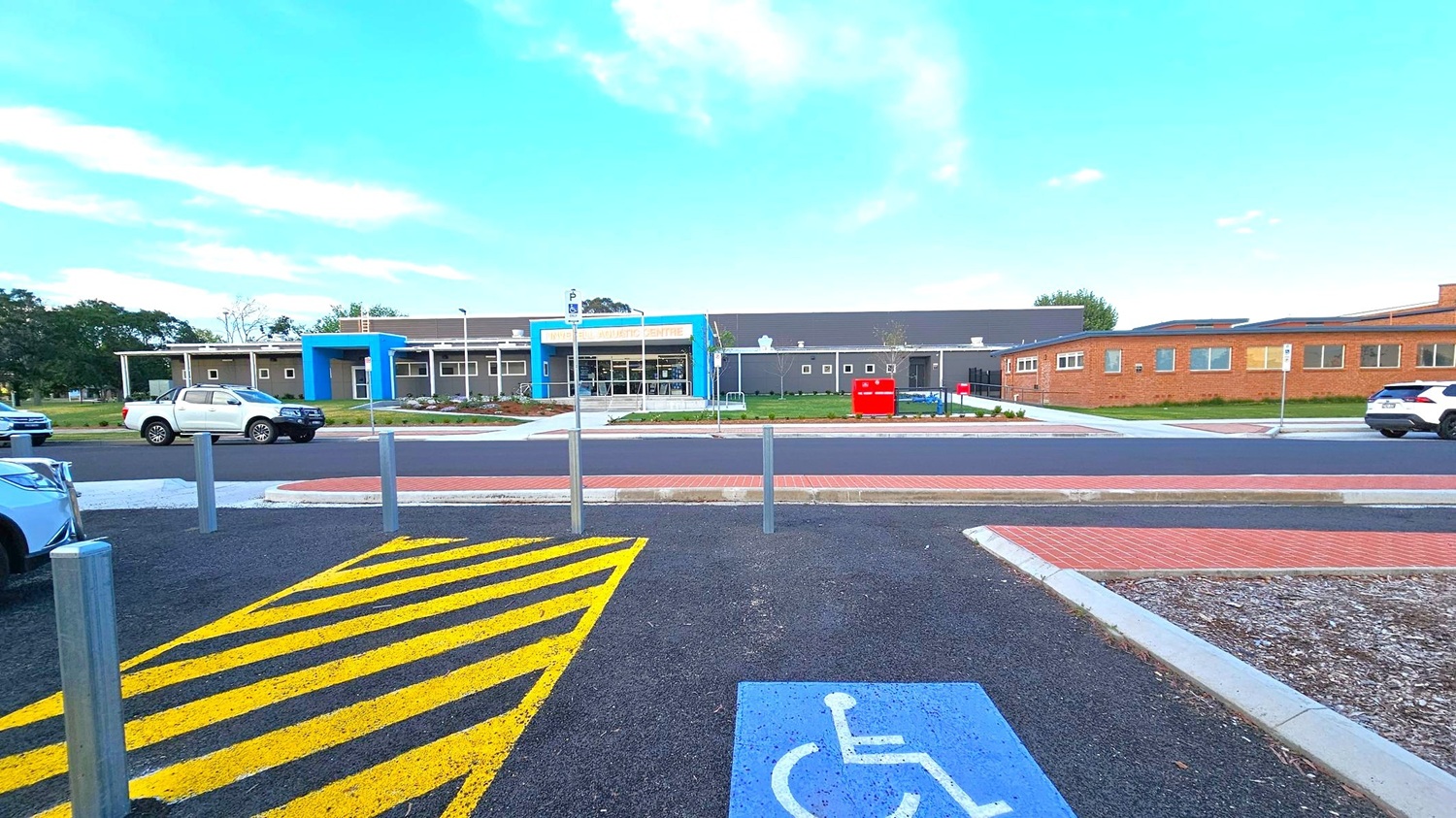 A building with a blue entrance and a sign above it is in the centre. The structure is surrounded by a paved area with a few parked cars, including a white pickup truck. In the foreground, there is a parking space marked with a blue disabled symbol and yellow diagonal lines. The ground is a mix of asphalt and brick paving. Trees and grass are visible in the background.