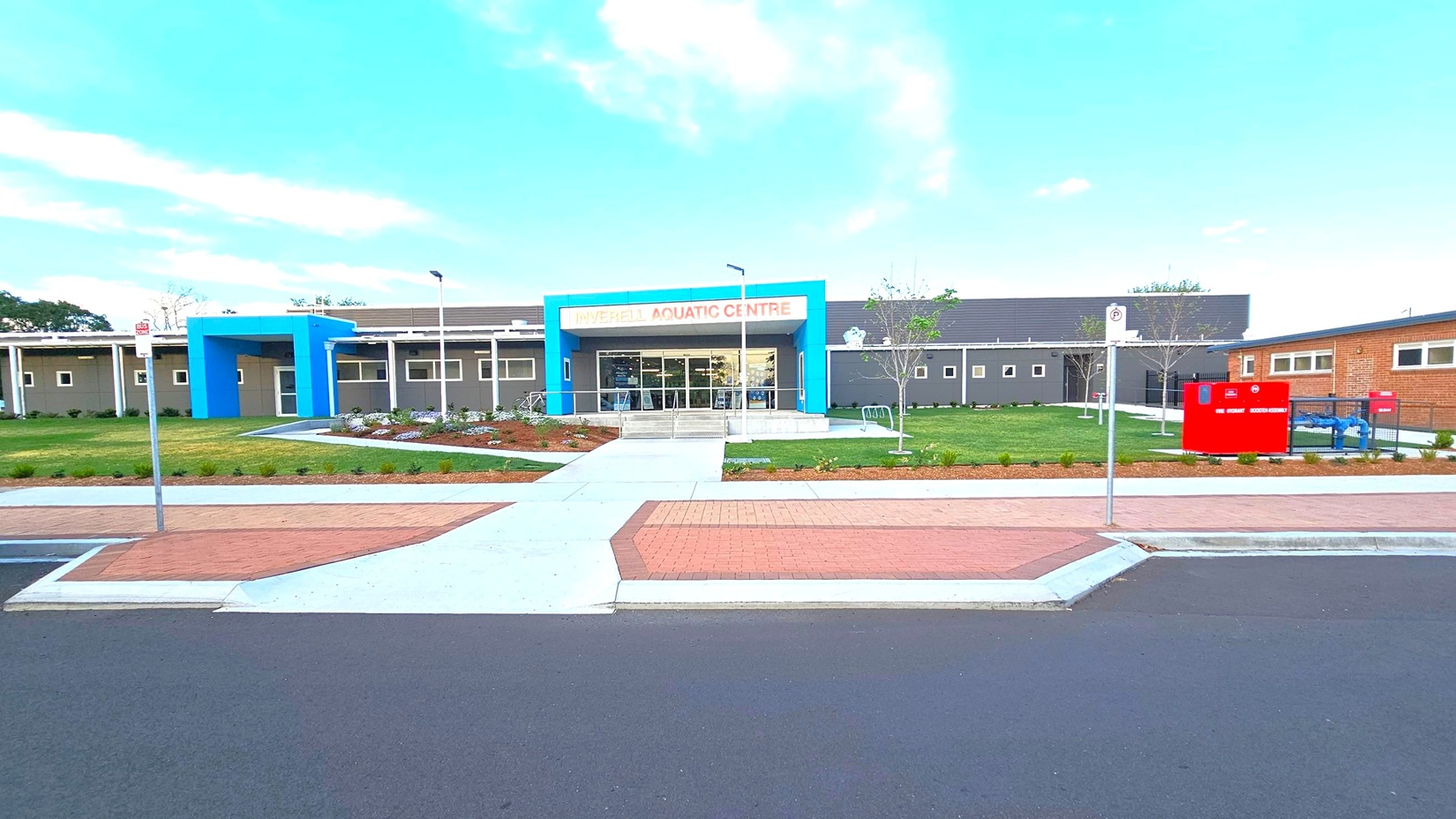 A modern building with a blue entrance and a sign reading "Inverell Aquatic Centre" in orange letters. The structure is surrounded by a well-maintained lawn and landscaped garden beds. A pathway leads from the street to the entrance. On the right, there is a red sign and some equipment. The foreground features a paved area and a road with a no-parking sign. The building has a flat roof and several small windows.