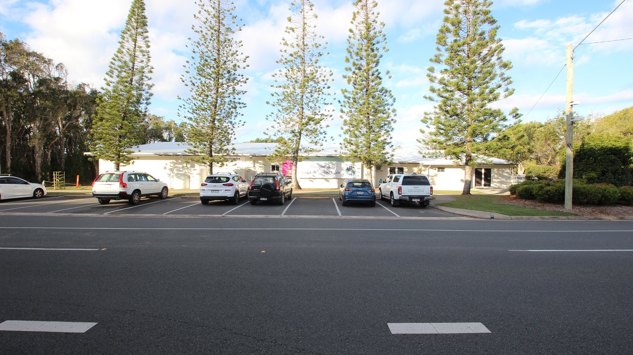 A row of parked cars is in front of a single-storey building with a light-coloured exterior. Tall, narrow trees are evenly spaced along the front of the building. The foreground features a multi-lane road with visible lane markings. On the left, there are more trees, while on the right, there is a grassy area with bushes and a utility pole.