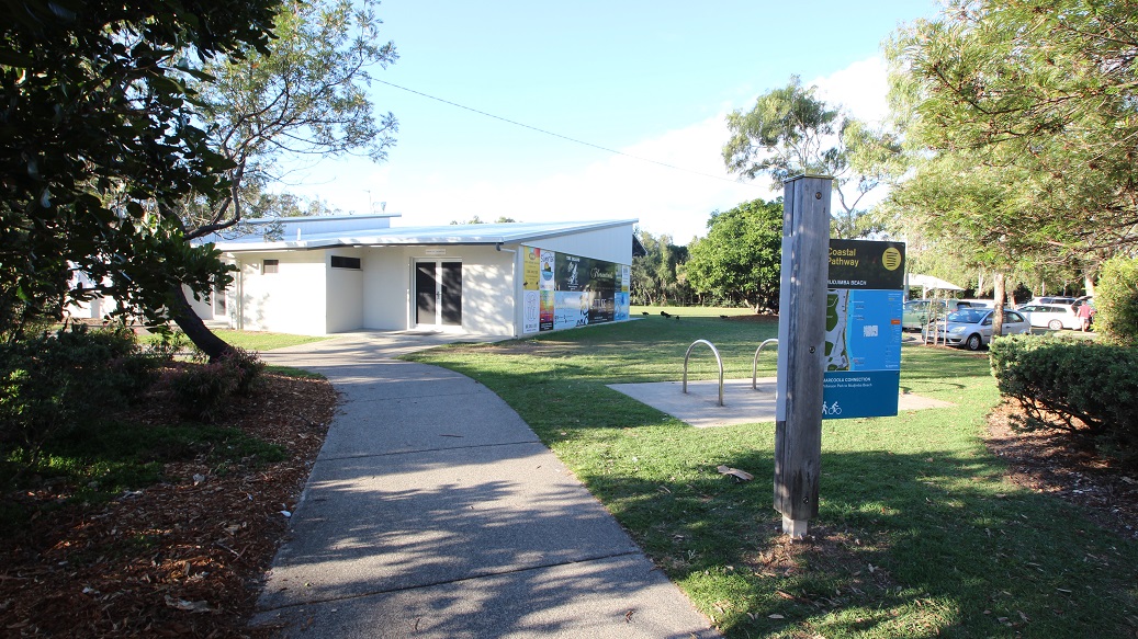 A pathway leads to a modern, single-storey building with large windows on the left. In the foreground, a signpost with a blue information board stands beside a bike rack. The area is surrounded by well-maintained grass and trees, with a few cars parked on the right. The scene is well-lit, suggesting daytime.