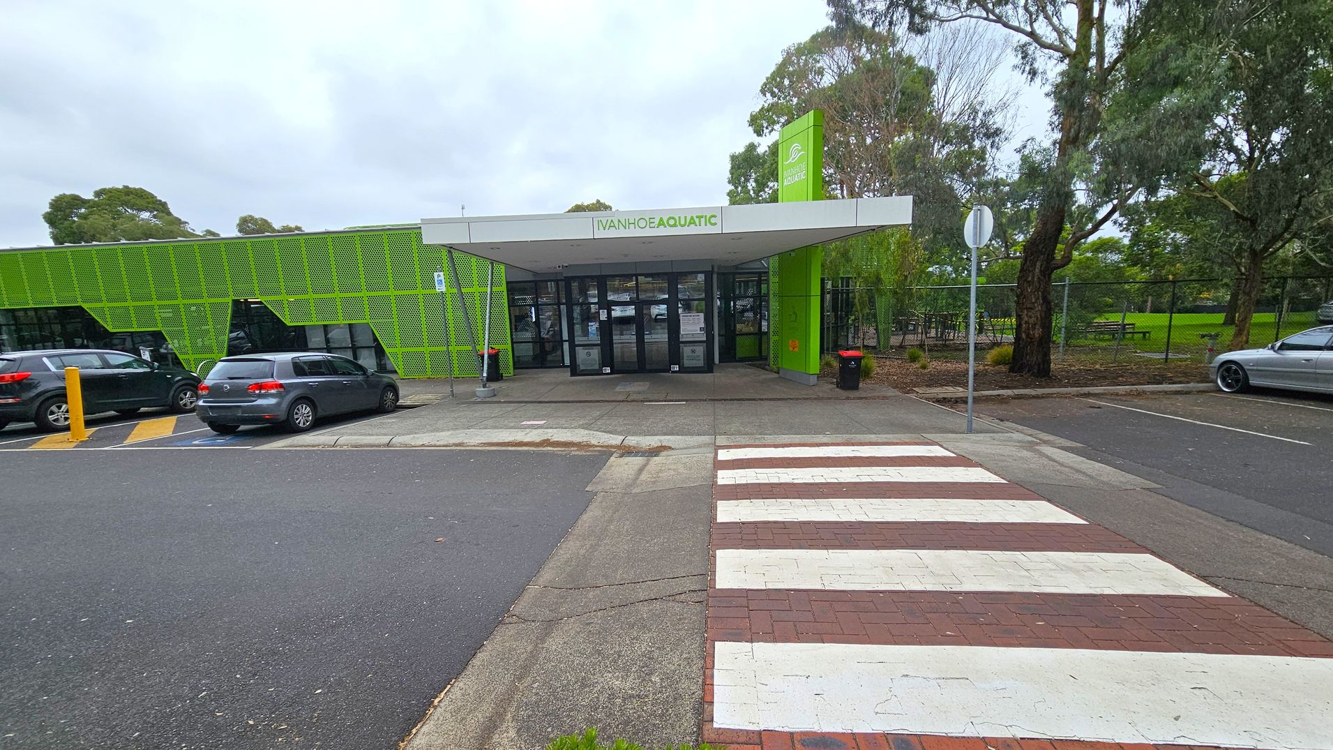 A building with a green facade and a sign reading "Ivanhoe Aquatic" above the entrance. The entrance has glass doors and a covered walkway. On the left, there are parked cars next to the building. On the right, there are trees and a grassy area with a picnic table. A zebra crossing leads to the entrance from the foreground, with a mix of asphalt and brick paving. A stop sign is visible near the trees.