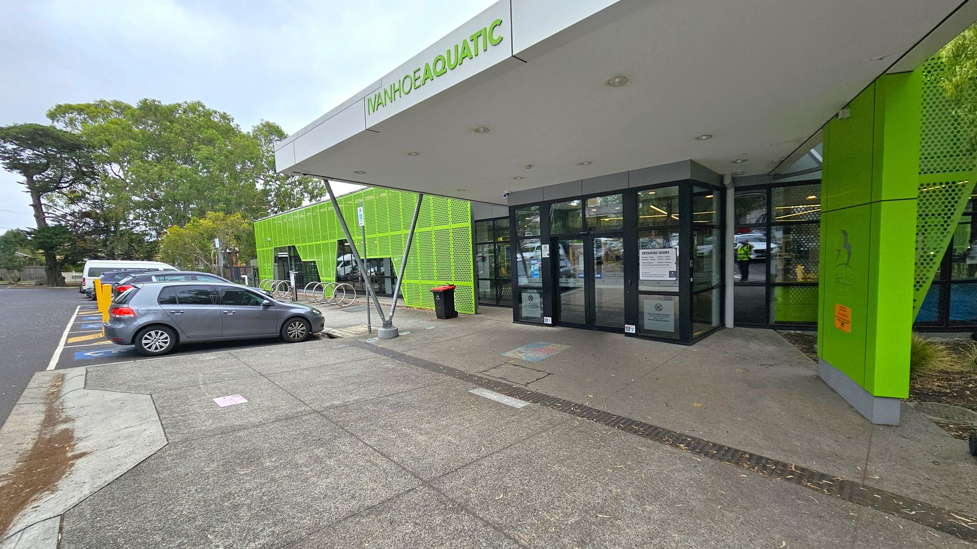The image shows the entrance to a building with a sign reading "Ivanhoe Aquatic" above the door. The building has a modern design with bright green panels. On the left, there is a car park with a grey car and a white van parked in designated spaces. A row of bicycle racks is visible near the entrance. The ground is paved with concrete, and there are trees in the background. The entrance has glass doors with notices displayed on them.