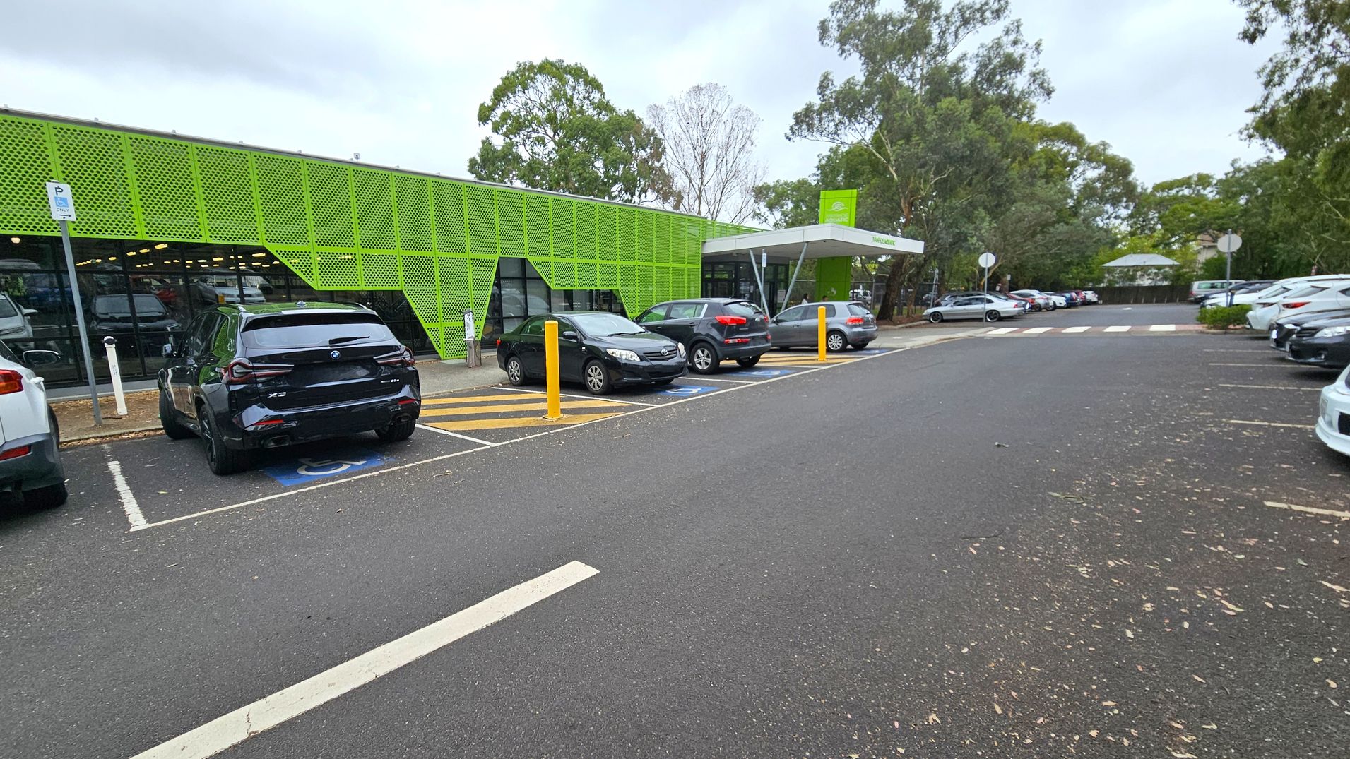A car park with several vehicles is in front of a building with a distinctive green perforated facade on the left. The building has large glass windows beneath the green panels. The car park has designated spaces, including a disabled parking spot marked in blue. Yellow bollards are present near the entrance. Trees and foliage surround the area, with more parked cars visible in the background. The ground is asphalt, and there are some scattered leaves.