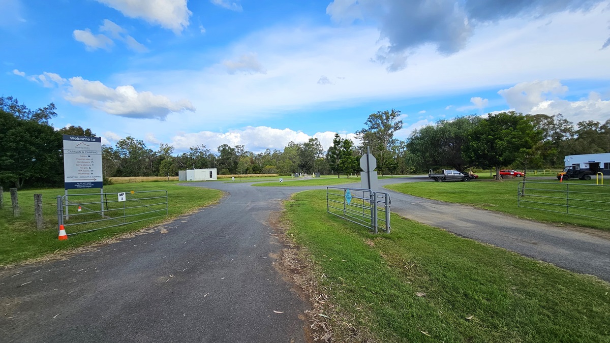 A paved road leads into a grassy area with a large sign on the left, next to a wire fence and an orange traffic cone. In the background, there are trees and a small white building. On the right, a few vehicles are parked near another building. The sky is partly cloudy.