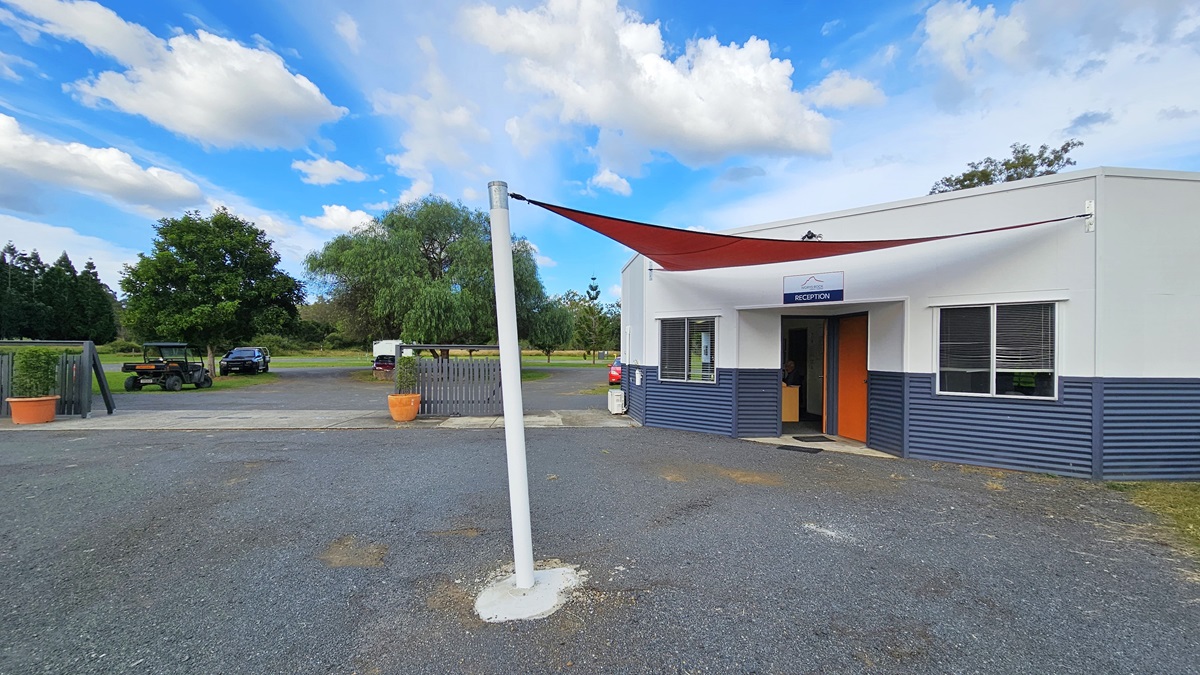 A small building with a sign reading "Reception" above the entrance is on the right. The building has a white and blue exterior with a red door. A red triangular shade sail is attached to a white pole in the foreground. On the left, there are trees and a few parked vehicles in the background. The ground is covered with gravel, and there are large potted plants near the entrance.