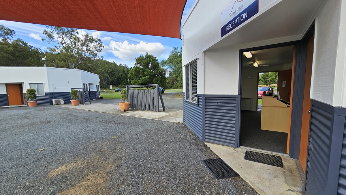 A gravel driveway leads to a building with a reception area on the right. The building has a grey and white exterior with a sign above the entrance. The door is open, revealing a lit interior with a desk. On the left, another building with a similar design is visible, along with potted plants. In the background, there are trees and a grassy area. A red shade cloth is partially visible above.