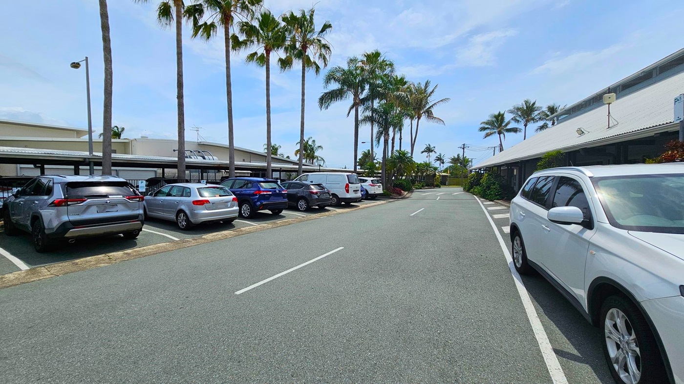 A car park with several vehicles on the left, including a silver SUV and a blue car. Tall palm trees line the area, providing shade. On the right, a white SUV is parked near a building with a sloped roof. The road in the centre is clear, with white lines marking the lanes. The background features more palm trees and a few low-rise buildings.