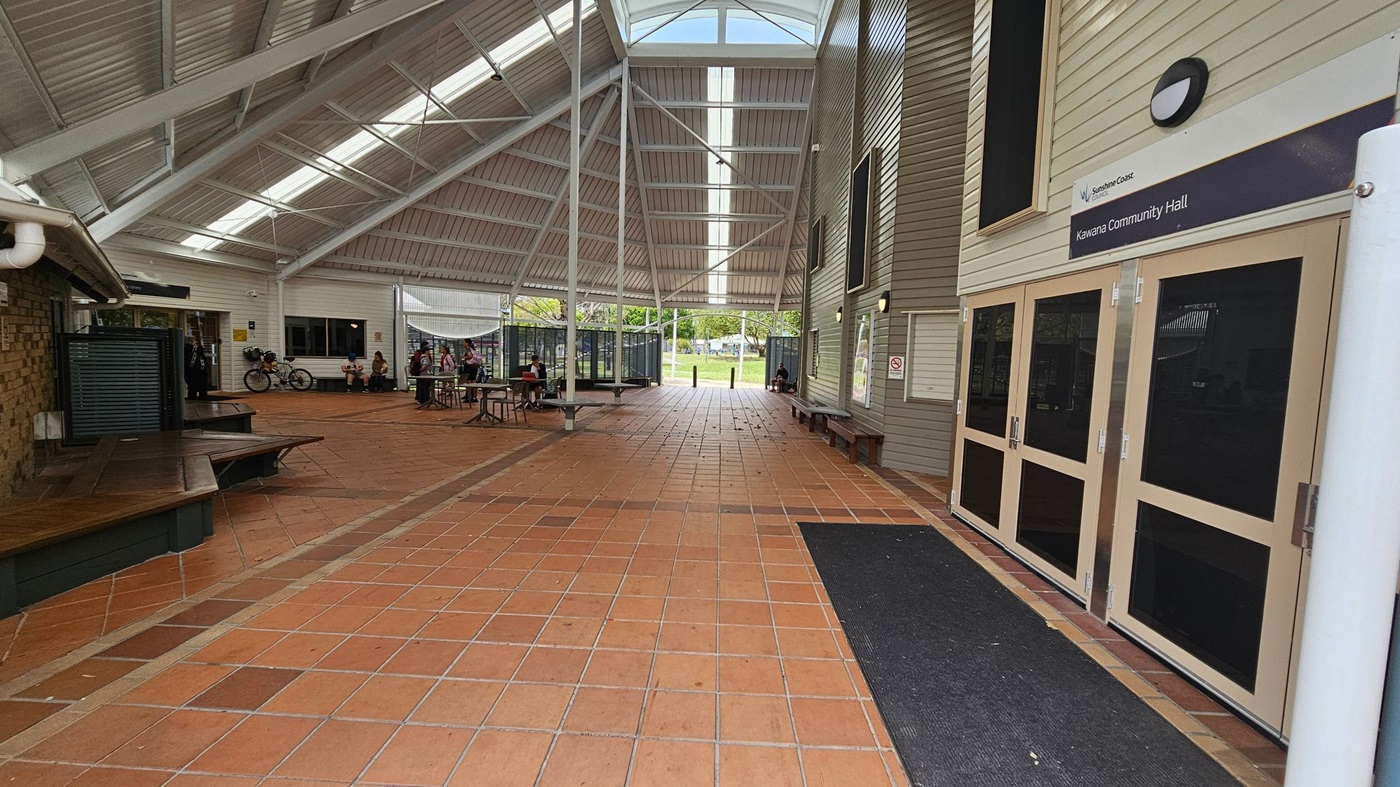 A spacious indoor area with a high, sloped ceiling featuring skylights. The floor is covered with terracotta tiles. On the right, there are doors labelled "Kawana Community Hall" and a wall with benches. On the left, a brick wall with a bench and a bicycle parked nearby. In the middle, several tables and chairs are scattered, with people sitting and conversing. The background shows an open area with greenery visible through the entrance.