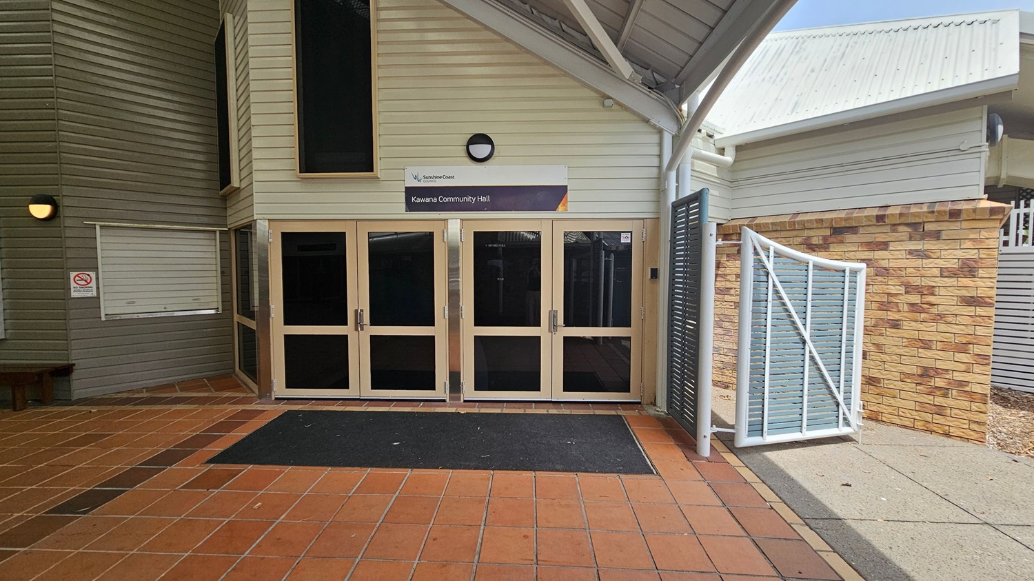 Entrance to a community hall with a sign above the doors reading "Kawana Community Hall." The building has light-coloured cladding and a sloped roof. The entrance features four glass doors with a black mat in front. To the left, there is a green wall with a small window and a "No Smoking" sign. On the right, a brick wall and a white gate with blue slats are visible. The floor is tiled in a reddish-brown pattern.