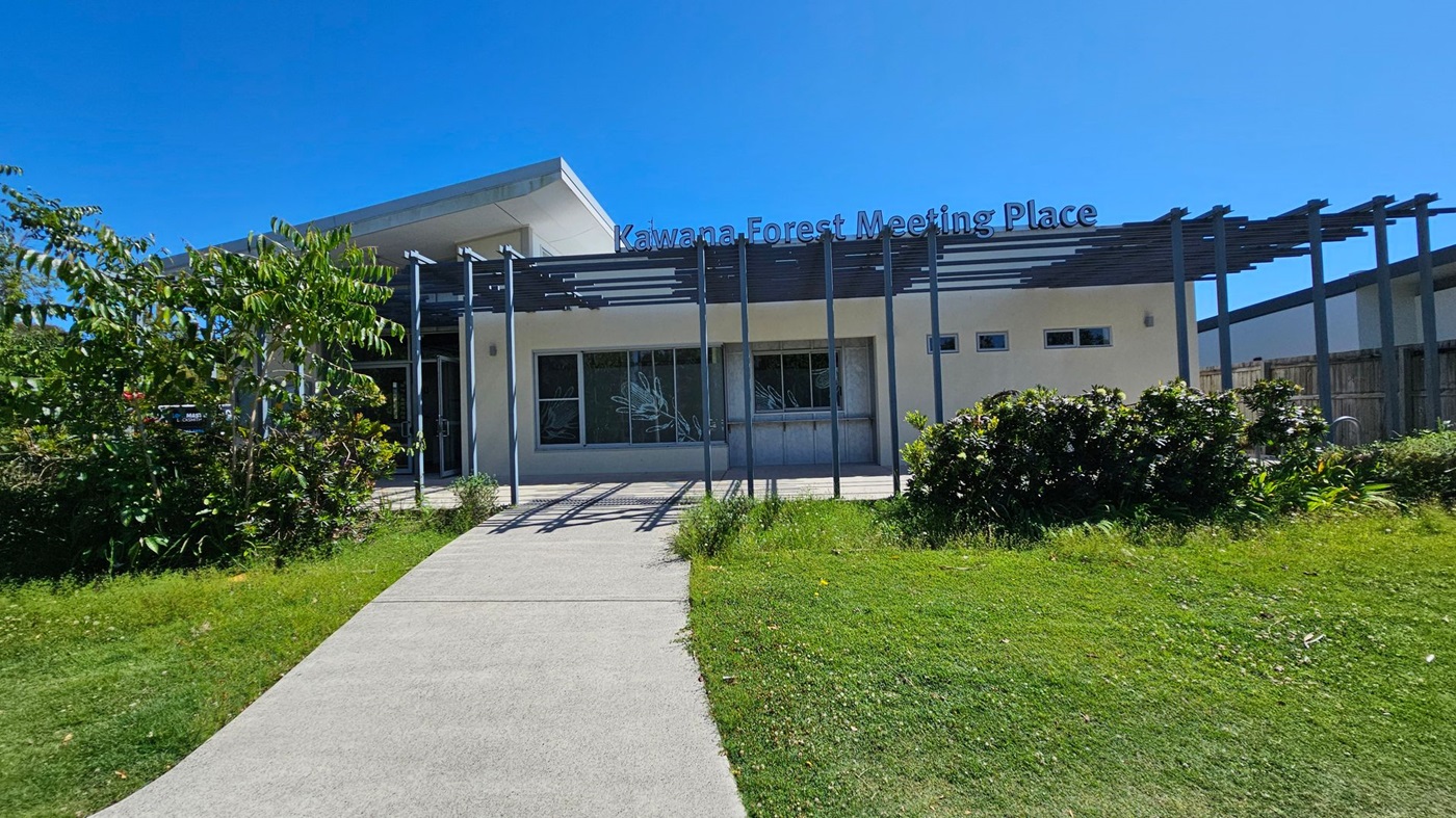 A modern building with a flat roof and a sign reading "Kawana Forest Meeting Place" on top. The structure features large windows and a pergola with vertical beams. A concrete path leads to the entrance, surrounded by well-maintained grass and shrubs. On the left, there are trees and more greenery, while the right side has additional plants and a wooden fence. The sky is clear and blue.