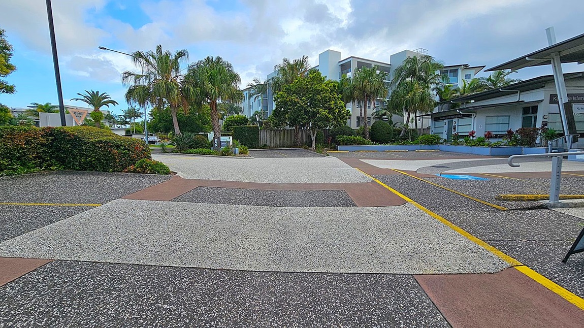 A street scene with a textured, multi-coloured pavement featuring grey, brown, and black sections. On the left, there are lush green bushes and palm trees, with a "Give Way" sign visible. In the centre, a modern white building is partially obscured by more palm trees and greenery. On the right, a building with a sloped roof and a sign is surrounded by landscaped plants. A metal railing is visible in the foreground.