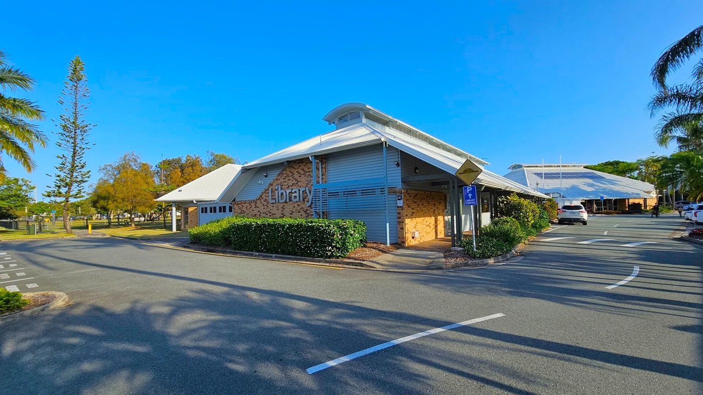 A library building with a modern design, featuring a sloped roof and a mix of brick and grey panelling. The word "Library" is prominently displayed on the side. In the foreground, there is a paved road with parking spaces and a sign indicating library parking. On the left, tall trees and a grassy area are visible. On the right, more parking spaces and a few cars are present, with palm trees lining the area. Shadows of trees are cast on the pavement.